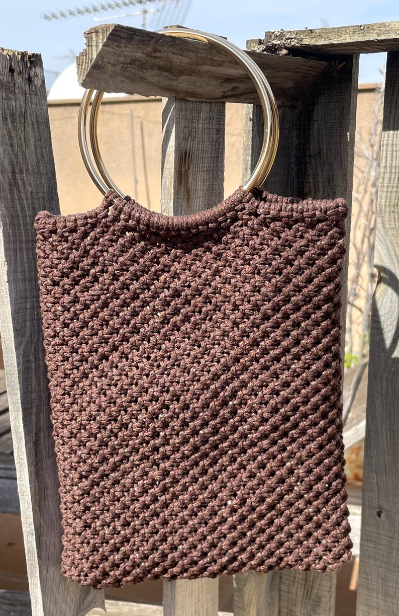 Pu&ograve; includere: Una borsa tote in macram&egrave; marrone con manici in metallo dorato. La borsa &egrave; rettangolare con una trama intrecciata. Le maniglie circolari sono attaccate alla parte superiore. La borsa &egrave; appesa a una staccionata di legno.
