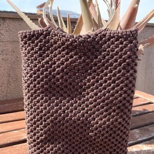 Pu&ograve; includere: Una borsa tote in macram&egrave; marrone con forma rettangolare e due manici in metallo argentato. La borsa &egrave; realizzata in un materiale tessuto con un motivo strutturato. La borsa &egrave; appoggiata su una superficie di legno.