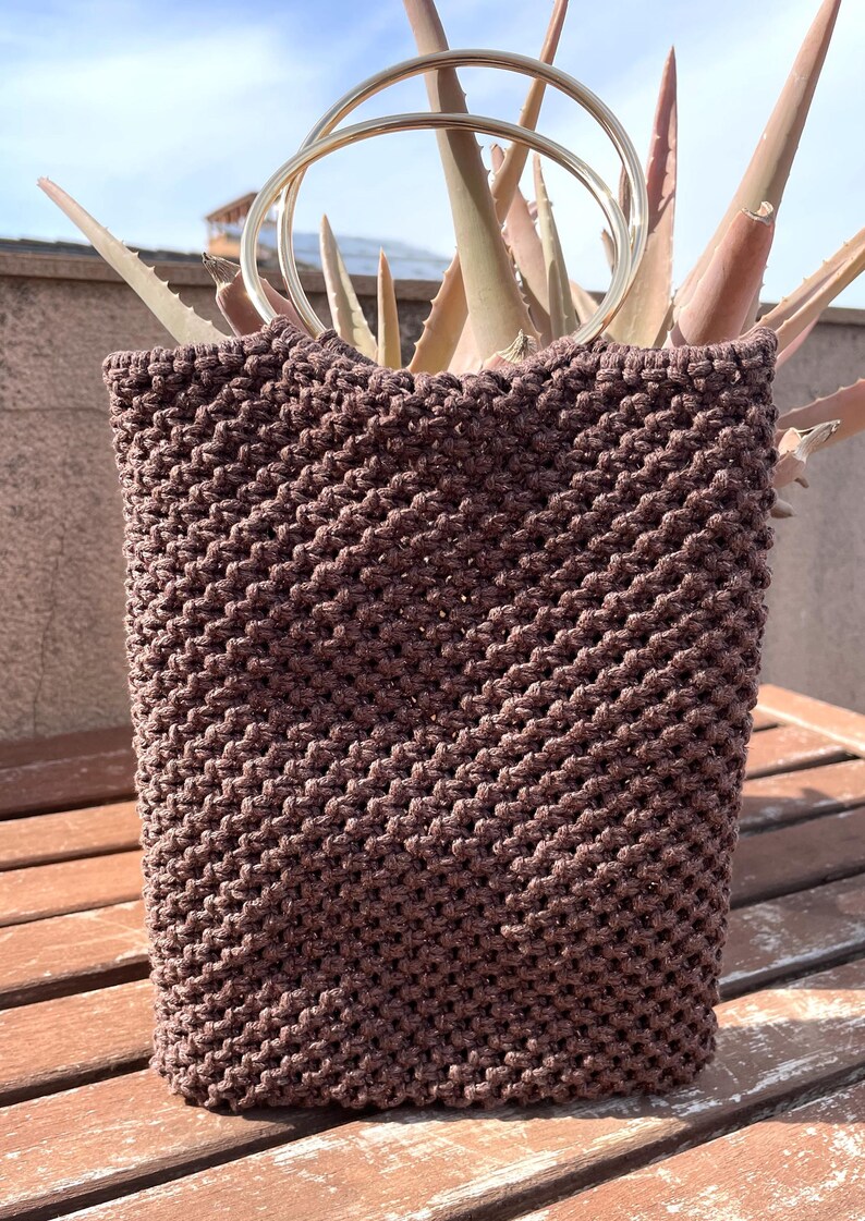Pu&ograve; includere: Una borsa tote in macram&egrave; marrone con forma rettangolare e due manici in metallo argentato. La borsa &egrave; realizzata in un materiale tessuto con un motivo strutturato. La borsa &egrave; appoggiata su una superficie di legno.