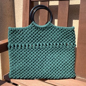 Puede incluir: Un bolso tote de macramé verde esmeralda hecho a mano con asas circulares negras. El bolso presenta un diseño tejido texturizado y se exhibe en una silla de madera. Este elegante bolso es perfecto para llevar lo esencial.