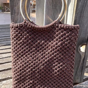 Pu&ograve; includere: Una borsa tote in macram&egrave; marrone con manici circolari color oro. La borsa &egrave; realizzata in un materiale a trama fitta e strutturata. La borsa &egrave; di forma rettangolare e sembra fatta a mano. La borsa &egrave; appoggiata su una superficie di legno.