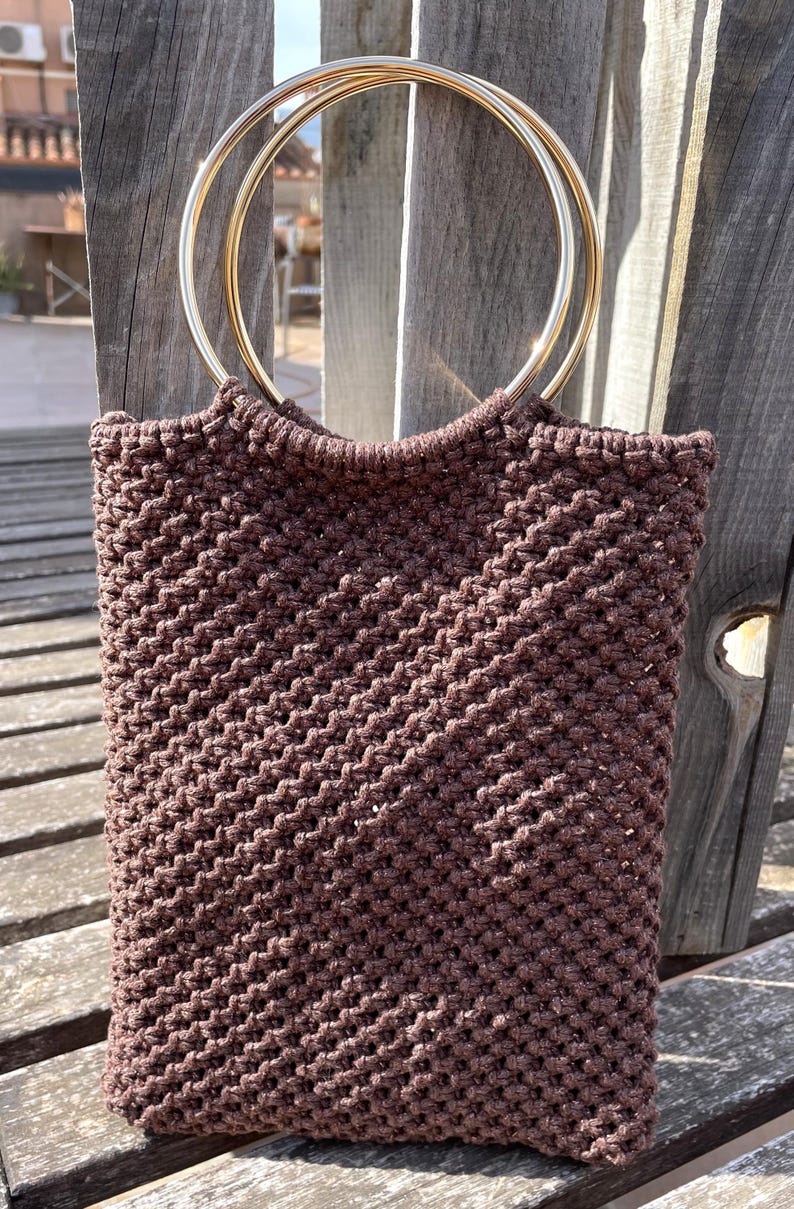 Pu&ograve; includere: Una borsa tote in macram&egrave; marrone con manici circolari color oro. La borsa &egrave; realizzata in un materiale a trama fitta e strutturata. La borsa &egrave; di forma rettangolare e sembra fatta a mano. La borsa &egrave; appoggiata su una superficie di legno.