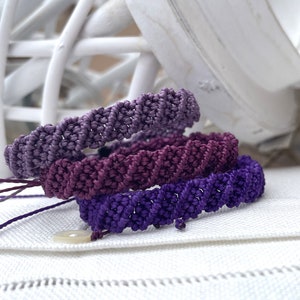May include: Three braided bracelets in shades of purple, lavender, and dark purple. The bracelets are stacked on top of each other and are made with a woven knot pattern.