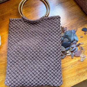 Pu&ograve; includere: Una borsa tote in macram&egrave; marrone con manici circolari color oro. La borsa &egrave; rettangolare e ha una trama intrecciata. Una collezione di vetro marino e ciottoli &egrave; accanto alla borsa su una superficie di legno.