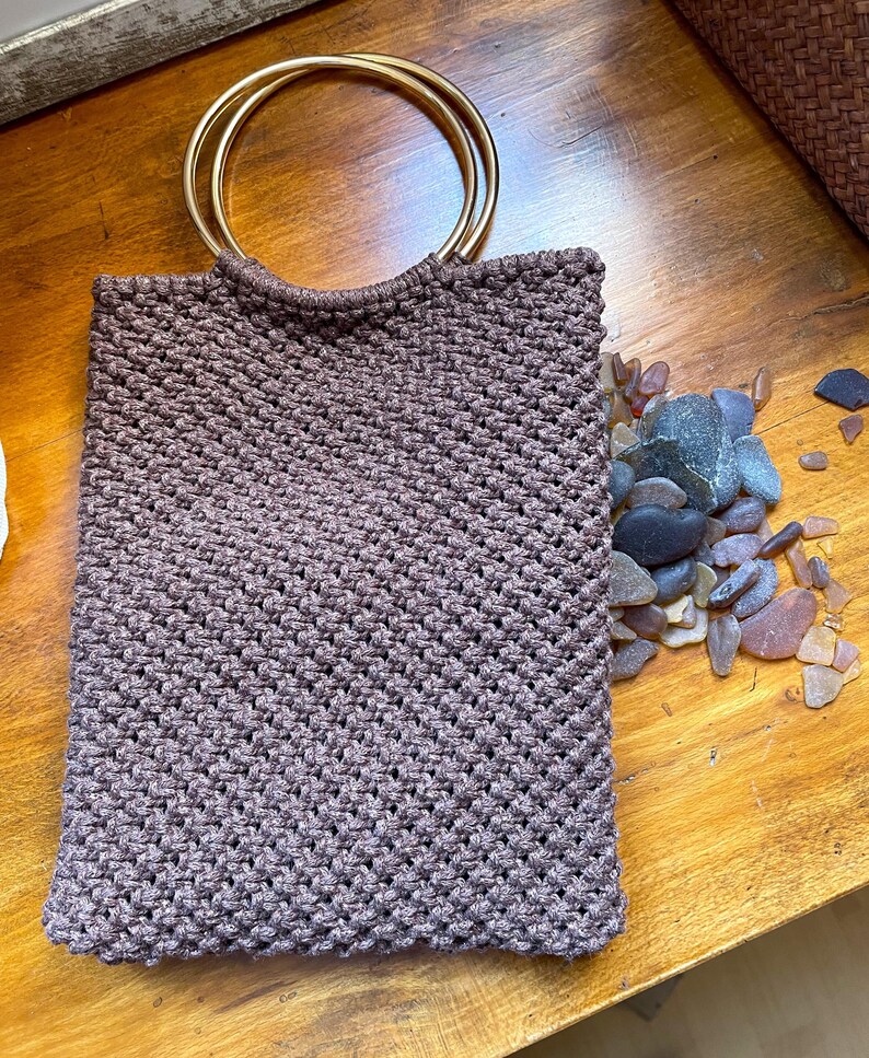 Pu&ograve; includere: Una borsa tote in macram&egrave; marrone con manici circolari color oro. La borsa &egrave; rettangolare e ha una trama intrecciata. Una collezione di vetro marino e ciottoli &egrave; accanto alla borsa su una superficie di legno.