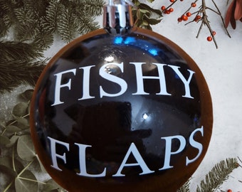 May include: A dark brown Christmas bauble with the words "FISHY FLAPS" in white block letters. The bauble has a silver metal cap and a loop for hanging. The background includes evergreen branches and red berries.