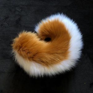 May include: A fluffy, circular hair scrunchie with a mix of brown and white faux fur. The scrunchie is a warm colour combination and has a hole in the centre. The accessory is designed to hold hair in place.