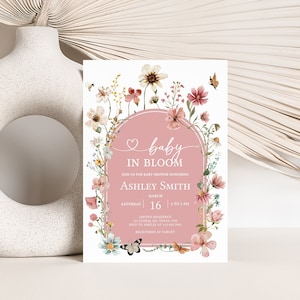 May include: A pink and white baby shower invitation with a floral wreath border. The text reads "baby in bloom" and includes the name Ashley Smith, the date March 16, and the time 2 to 3 PM.