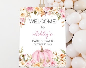 Puede incluir: Un letrero de bienvenida para el baby shower de Ashley, con una calabaza rosa y detalles florales. El letrero está sobre un caballete, con globos blancos en el fondo. El texto dice "Bienvenida al Baby Shower de Ashley 28 de octubre de 2023."