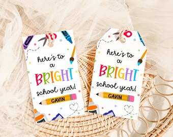 Op de afbeelding: Twee witte cadeaulabels met een schoolthema. Elk label heeft de tekst "here's to a BRIGHT school year!" in kleurrijke letters, samen met illustraties van schoolbenodigdheden zoals scharen, kleurpotloden en een potlood met de naam "GAVIN".