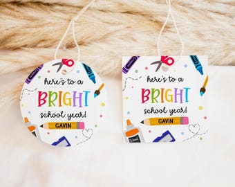 Op de afbeelding: Twee schoolthema-ornamenten met de tekst "here's to a BRIGHT school year!" en de naam "GAVIN". Eén is rond, de andere vierkant. Beide hebben kleurrijke illustraties van schoolbenodigdheden.
