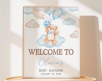 Peut inclure: Affiche de bienvenue pour la fête prénatale d'Olivia avec un ours en peluche brun tenant un nœud bleu et flottant sur des nuages bleus et blancs avec des ballons. Le texte est en brun et la date est le 25 janvier 20XX.