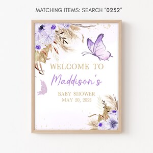 May include: A white sign with a wooden frame, featuring a purple butterfly, floral accents, and the text "WELCOME TO Maddison's BABY SHOWER MAY 20, 2023".