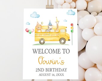 May include: A welcome sign for a 2nd birthday party. The sign features a yellow school bus with animal characters, balloons, and the text "Welcome to Gavin's 2nd Birthday August 16, 2XXX". The sign is displayed on a wooden easel.