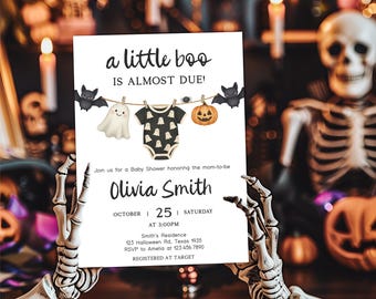 Può includere: Un invito per un baby shower a tema Halloween con il testo "a little boo is almost due!". L'invito presenta illustrazioni di un fantasma, vestiti per bambini, una zucca e pipistrelli. L'invito è tenuto da una mano di scheletro.