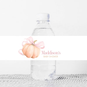 Puede incluir: Una botella de agua de plástico transparente con una etiqueta blanca que presenta una calabaza de acuarela con un lazo rosa. La etiqueta también incluye el texto "Maddison's BABY SHOWER" en rosa.