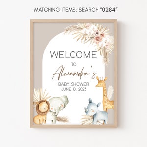 May include: A welcome sign for a baby shower with a light brown background and a white circle in the center. The text "WELCOME TO Alexandra's BABY SHOWER JUNE 10, 2023" is in the circle. There are illustrations of a lion, elephant, rhinoceros, and giraffe around the circle.