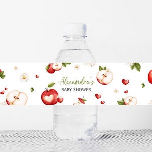 Op de afbeelding: Een waterfles met een aangepast etiket voor een babyshower. Het etiket bevat aquarel illustraties van rode appels, appelschijfjes, madeliefjes en rode harten op een witte achtergrond. De tekst "Alexandra's BABY SHOWER" is in groene letters geschreven.