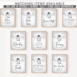 May include: A collection of framed signs with a winter theme, featuring a cartoon penguin. The signs have text such as "Babies are Sweet", "Books & Gifts", "Diaper Raffle", and "Don't Say Baby". The signs are framed in light wood.