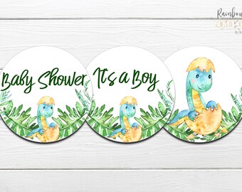 Könnte beinhalten: Drei runde Aufkleber mit einem Baby-Dinosaurier-Design. Der erste Aufkleber sagt "Baby Shower", der zweite Aufkleber sagt "It's a Boy" und der dritte Aufkleber zeigt einen blauen Dinosaurier, der aus einem Ei schlüpft.