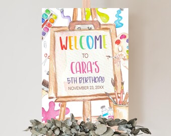 May include: Welcome to Cara's 5th Birthday party sign with a watercolor illustration of an easel, paintbrushes, paint tubes, and a palette. The sign is on a wooden easel.