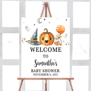 May include: A white sign on a wooden easel with a Halloween-themed baby shower design. The sign features a smiling pumpkin, witch hat, and the text "WELCOME TO Samantha's BABY SHOWER NOVEMBER 6, 2021."