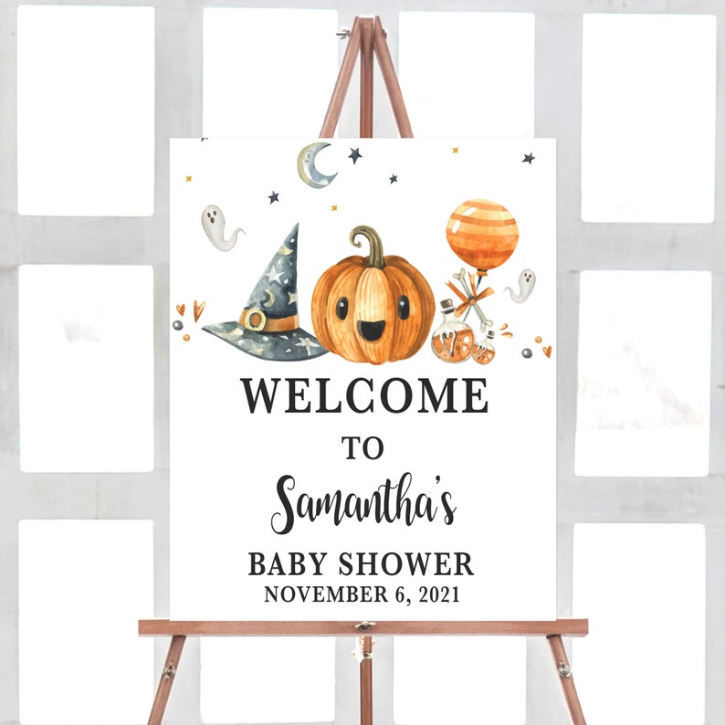 May include: A white sign on a wooden easel with a Halloween-themed baby shower design. The sign features a smiling pumpkin, witch hat, and the text "WELCOME TO Samantha's BABY SHOWER NOVEMBER 6, 2021."