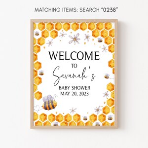 May include: A welcome sign for a baby shower with a honeycomb pattern and a bee illustration. The sign says "Welcome to Savannah's Baby Shower May 20, 2023".