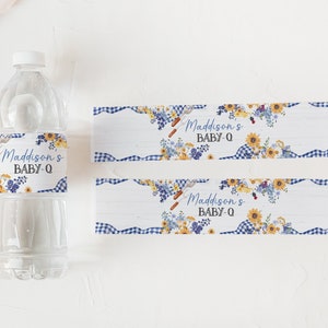 Puede incluir: Dos etiquetas con estampado de cuadros azules y blancos con un dise&ntilde;o floral y el texto "Maddison's Baby-Q" impreso en ellas. Las etiquetas est&aacute;n dise&ntilde;adas para botellas de agua.