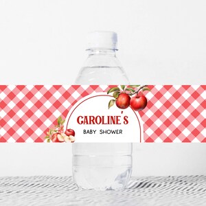 Op de afbeelding: Een waterfles met een aangepast etiket voor een babyshower. Het etiket heeft een rood-wit ruitpatroon met de tekst "CAROLINE'S BABY SHOWER" in een cirkel. Waterverf appels en bladeren versieren het etiket.