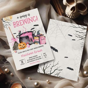 A Halloween-themed baby shower invitation featuring a witch's broom, a cauldron, and bats, along with a skull and spider web design.