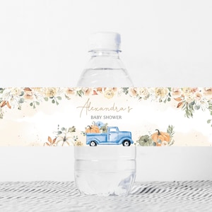 Puede incluir: Una botella de agua con una etiqueta decorativa. La etiqueta presenta un camión azul que transporta calabazas, con el texto "Alexandra's Baby Shower" en letras doradas. El diseño incluye elementos florales y de follaje en colores otoñales.