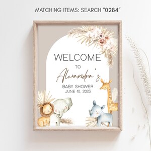 May include: A welcome sign for a baby shower with a light brown wooden frame. The sign has a white background with a light brown arch and a light brown welcome sign with the text "Welcome to Alexandra's Baby Shower June 10, 2023". The sign has illustrations of a lion, elephant, rhinoceros, and giraffe.