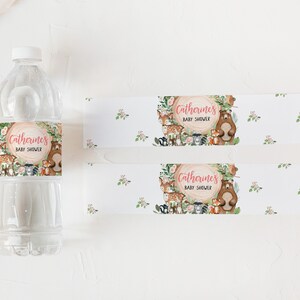 May include: Water bottle labels with a woodland theme. The labels feature a brown tree trunk with pink text that reads "Catherine's Baby Shower" surrounded by woodland animals including a bear, fox, deer, and squirrel. The labels are printed on white paper with a subtle floral pattern.