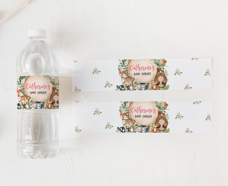 May include: Water bottle labels with a woodland theme. The labels feature a brown tree trunk with pink text that reads "Catherine's Baby Shower" surrounded by woodland animals including a bear, fox, deer, and squirrel. The labels are printed on white paper with a subtle floral pattern.
