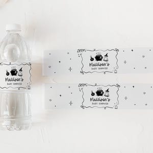 A clear plastic bottle and two white labels with black text and illustrations, likely for a baby shower, are placed on a white surface.