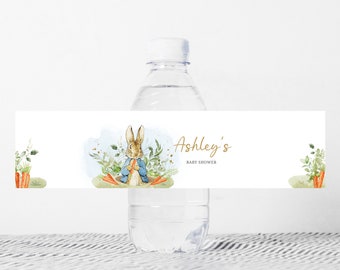 May include: Water bottle label featuring a watercolour illustration of Peter Rabbit holding a carrot. The label is white with green foliage and carrots. The text on the label reads "Ashley's Baby Shower".
