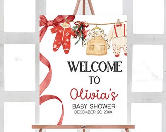 May include: A baby shower welcome sign on an easel. The sign features the text "Welcome to Olivia's Baby Shower" in black and red. Watercolor illustrations of baby clothes and a red ribbon adorn the top.