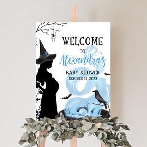 Op de afbeelding: Een welkomstbord voor een babyshower met een silhouet van een zwangere heks. De tekst "Welcome to Alexandra's Baby Shower October 14, 2023" wordt benadrukt met blauwe en zwarte elementen. Een kraai en vleermuizen zijn ook afgebeeld.