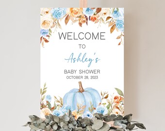 May include: A baby shower welcome sign on an easel. The sign features floral accents in shades of blue, orange, and cream. The text reads "Welcome to Ashley's Baby Shower October 28, 2023" with a blue pumpkin illustration.