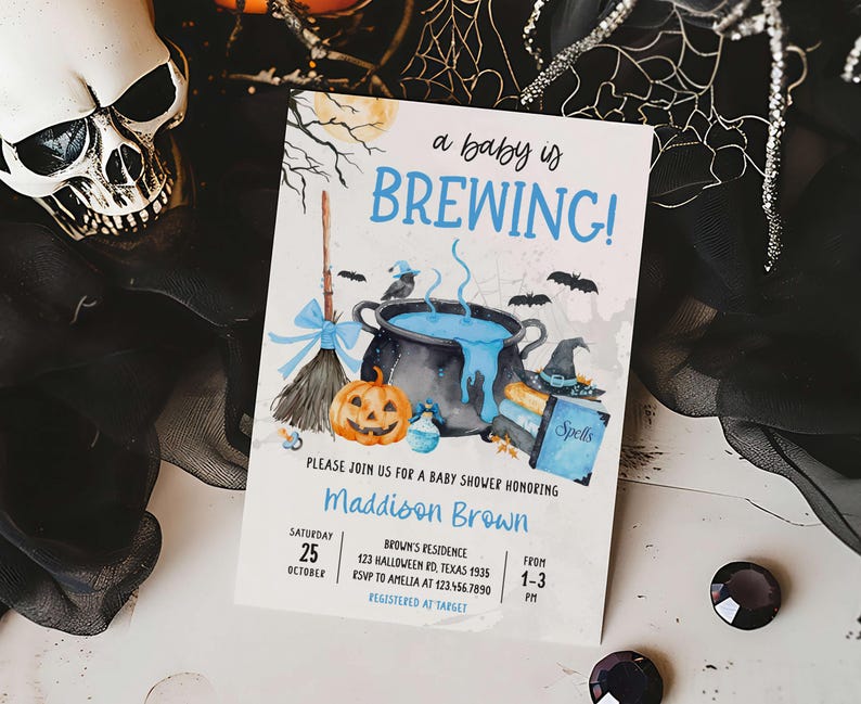 A Halloween-themed baby shower invitation featuring a skull, broom, cauldron, and pumpkins, with the text "a baby is BREWING!"