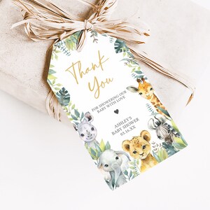 Puede incluir: Etiqueta de agradecimiento con tem&aacute;tica de la selva. Presenta un fondo blanco con letras doradas que dicen "Gracias" y "Por duchar a nuestro beb&eacute; con amor". Hay ilustraciones de una jirafa, un rinoceronte, un le&oacute;n, un elefante y una cebra rodeados de hojas verdes.