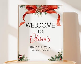 May include: A framed baby shower sign with a watercolour design. The sign reads "WELCOME TO Olivia's BABY SHOWER" in black and red text. A red bow and greenery decorate the top and bottom corners.