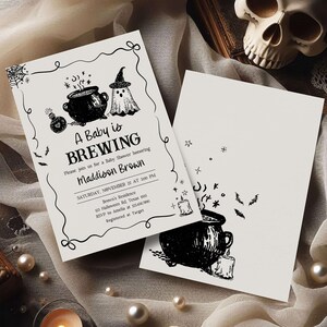 A Halloween-themed baby shower invitation featuring a black and white design with illustrations of a baby, a witch, and a cauldron.