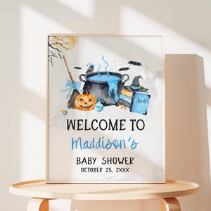 The image is a framed welcome sign for a baby shower, featuring illustrations of Halloween-themed items such as a cauldron, broomstick, jack-o-lantern, and bats.