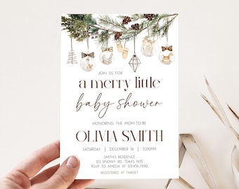 May include: A baby shower invitation with the text "a merry little baby shower" in brown script. The invitation features watercolor illustrations of baby-themed ornaments and pine branches. The invitation is held by a hand.
