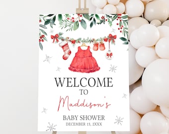 May include: A baby shower welcome sign on an easel. The sign features a red dress, stocking, and ornaments hanging from a garland. The text reads "WELCOME TO Maddison's BABY SHOWER DECEMBER 13, 2XXX". White balloons are in the background.