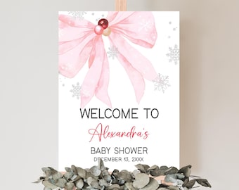 May include: A baby shower welcome sign on an easel. The sign features a large pink bow with snowflake accents and the words "Welcome to Alexandra's Baby Shower" in black and red text. Decorative greenery is at the base.