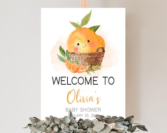 Puede incluir: Un letrero de bienvenida para un baby shower con una ilustración de acuarela de naranjas en una cesta. El letrero dice "Bienvenida a la fiesta de bienvenida de Olivia" con la fecha del 25 de enero de 2024. El letrero se muestra en un caballete de madera.