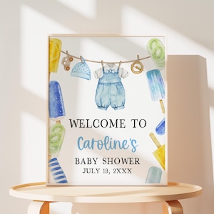 May include: A welcome sign for a baby shower with a blue and white baby outfit, popsicles, and the text "Welcome to Caroline's Baby Shower July 19, 2XXX"